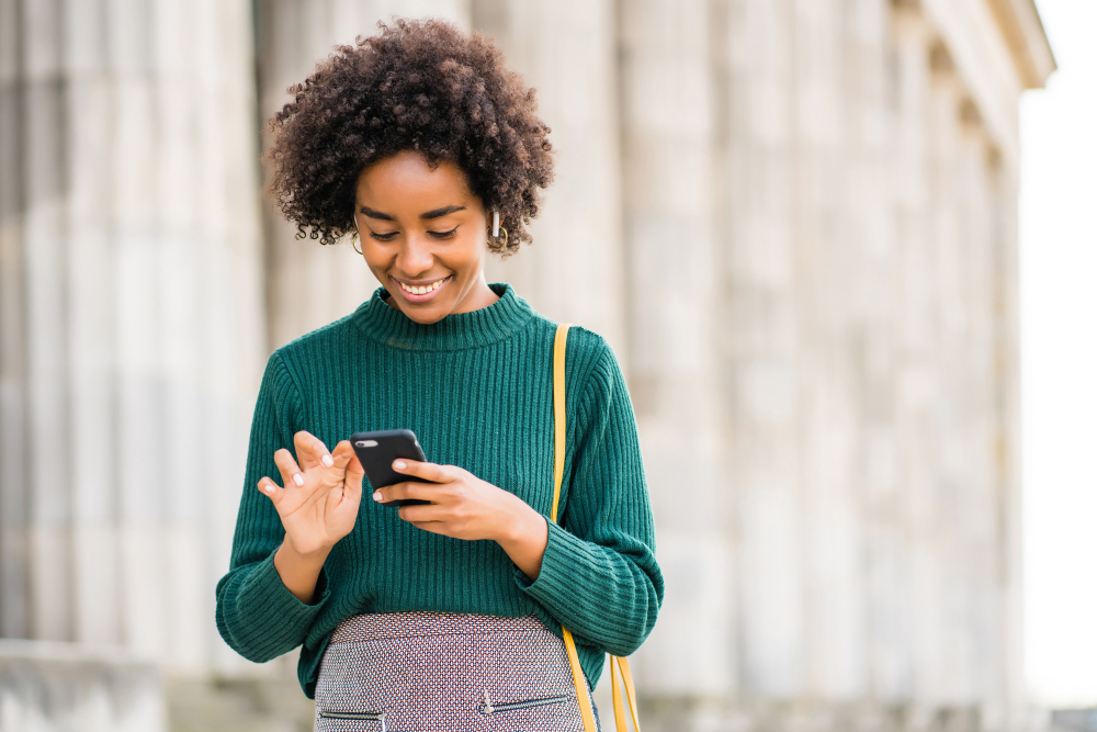 Mulher sorrindo enquanto usa celular ao ar livre, simbolizando os tipos de benefícios e qualidade de vida para colaboradores.